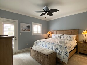 Un dormitorio con cama, dos lámparas de noche, ventilador de techo, banca de mimbre y una puerta con persianas.