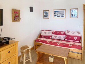 Um quarto aconchegante com uma cama-sofá de madeira decorada com motivos de coração, uma mesa de madeira, um banquinho pequeno e uma televisão sobre um móvel de madeira.