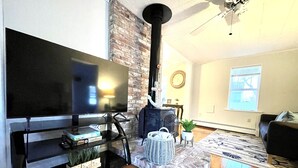 A living room with a flat-screen TV, a brick wall, a black stove, a potted plant, and a wooden floor. - Swan River Cottage - Near Beach, Water View, Kayak, (Dennis)