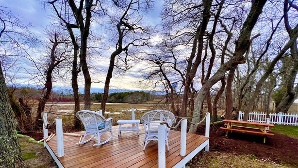 A wooden deck with white wicker chairs and a bench, surrounded by trees and a white fence. - Swan River Cottage - Near Beach, Water View, Kayak, (Dennis)