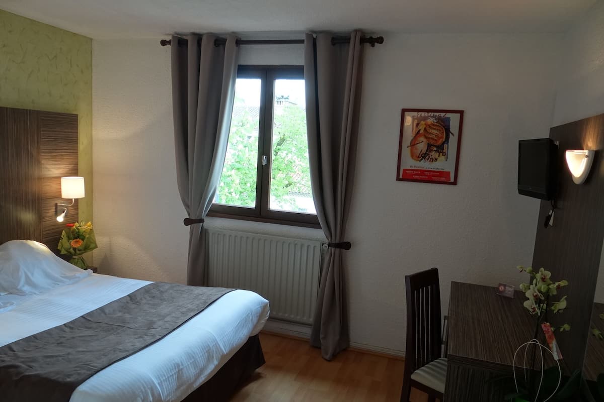 A hotel room with a bed, a desk, a chair, a radiator, a window with curtains, a wall-mounted TV, and a poster on the wall.