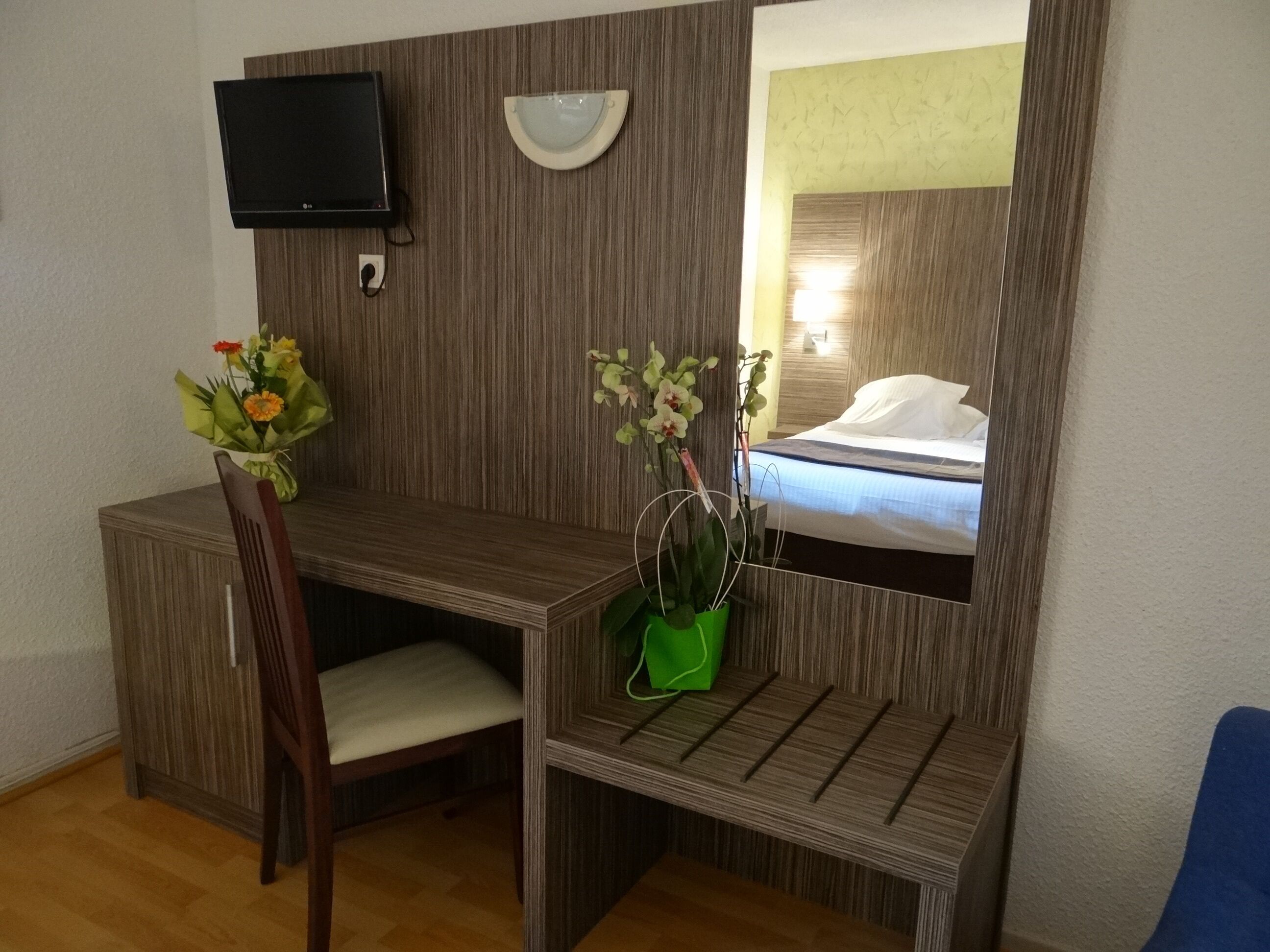 Quarto de hotel com escrivaninha de madeira, uma cadeira, televisão fixada na parede, mesa de cabeceira com um vaso verde e uma cama com travesseiro branco.