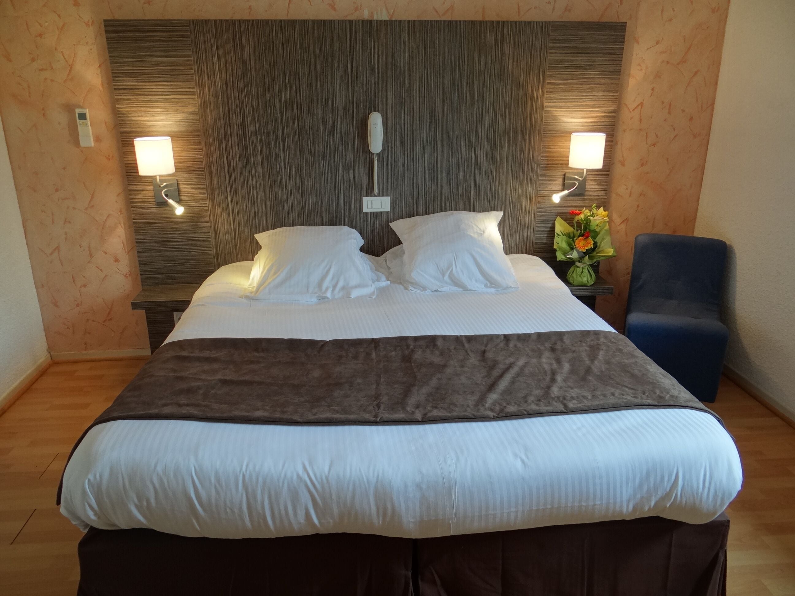 A neatly made bed with white linens and a brown throw blanket, flanked by two wall-mounted lamps.