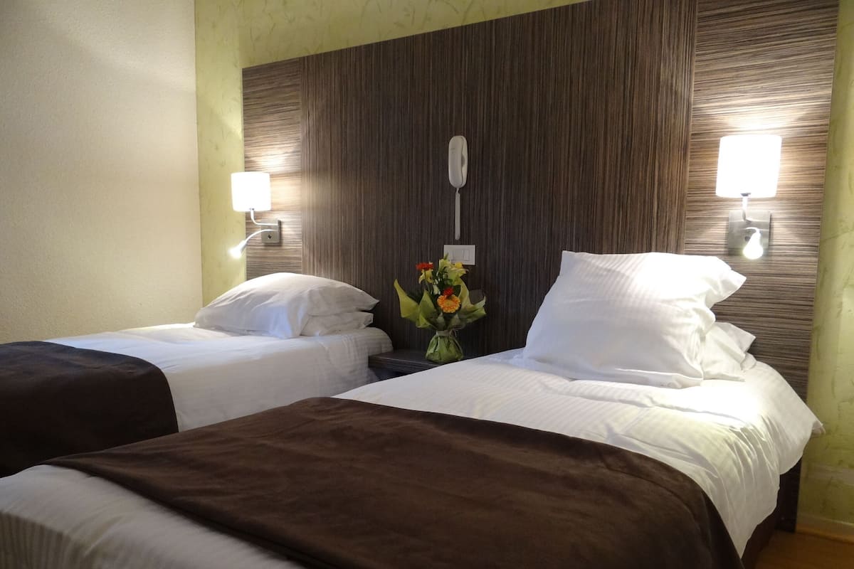 A hotel room with two beds, a nightstand with a lamp, a wall-mounted light, and a vase with flowers.