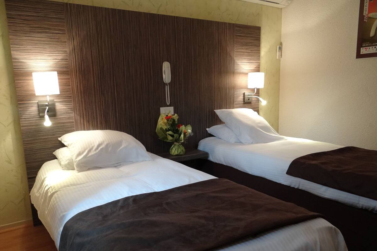 A hotel room with two beds, each with white bedding and a brown throw blanket. There is a nightstand between the beds with a lamp and a vase of flowers. The walls are covered with a wood panel design, and there are two wall-mounted lamps on either side of the room.