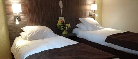 Habitación de hotel con dos camas, cada una con ropa blanca y una manta marrón. Entre las camas hay una mesita de noche con una lámpara y un florero. Las paredes tienen un diseño de paneles de madera, y hay dos lámparas de pared a cada lado de la habitación.