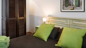 Habitación de hotel con cama, mesita de noche con lámpara, un cuadro en la pared y un juego de persianas de madera.