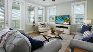 A living room with a ceiling fan, a flat-screen TV, and a view of a beach through the windows. - Seas The Day-Front Row-Beach Front-Next to Beach Club-3BD/2BA. Sleeps 8 (Galveston)