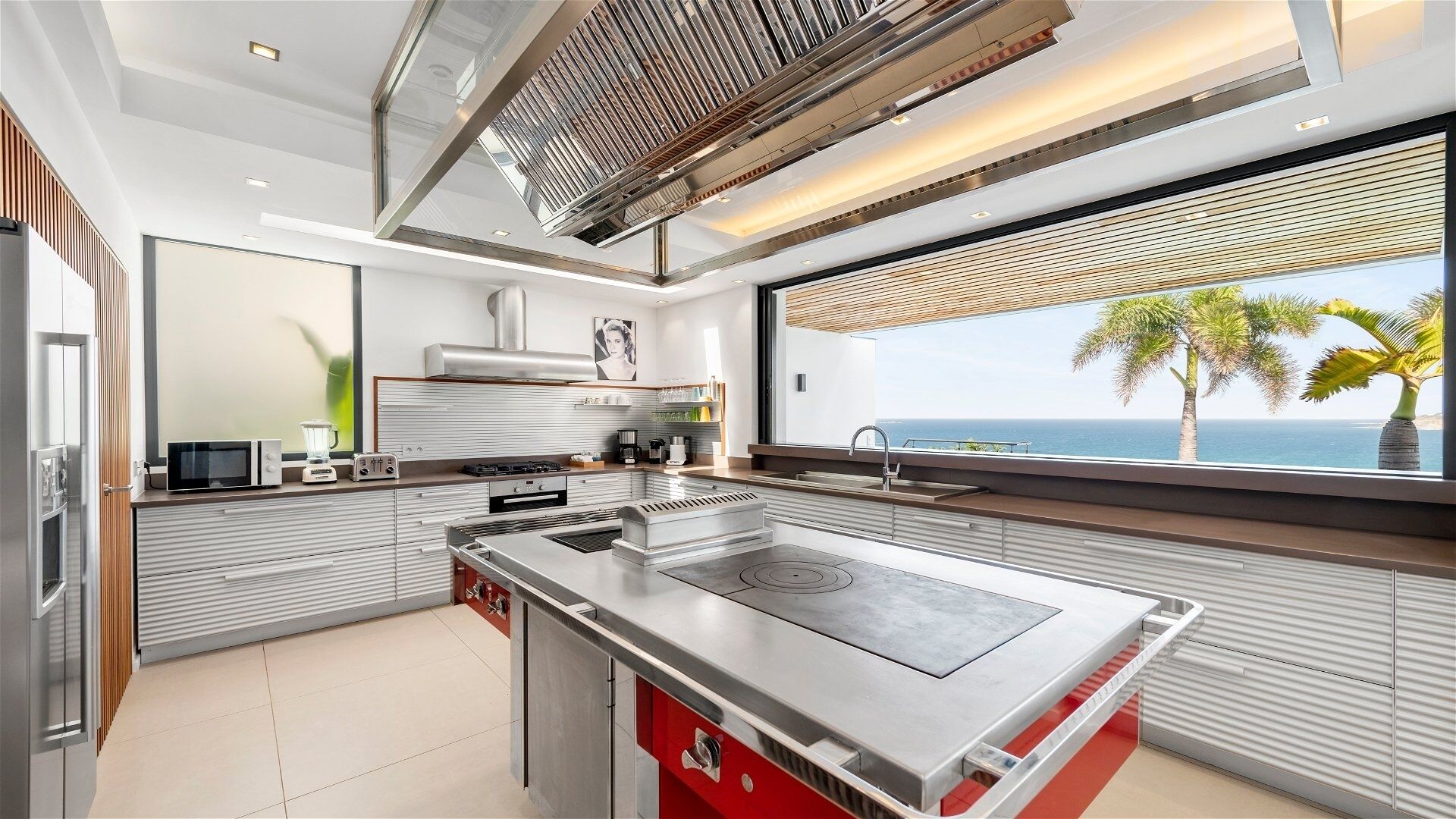 Une cuisine moderne dotée d’un îlot central, d’une grande fenêtre offrant une vue sur l’océan et d’un mur d’accent rouge.