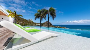 Een overloopzwembad met witte en groene ligstoelen, een houten terras en uitzicht op de oceaan en heuvels.