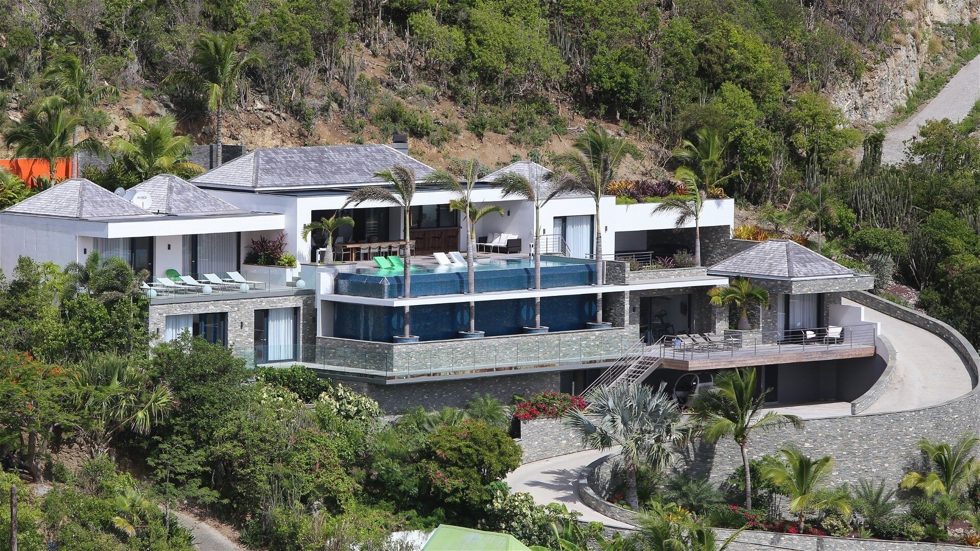A modern multi-story house with a rooftop infinity pool, surrounded by lush greenery and a hillside backdrop.