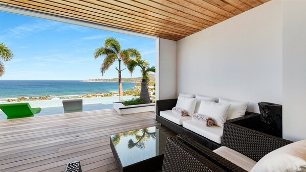 Une pièce moderne avec un plafond en bois, une table en verre, un canapé en osier avec des coussins blancs, et une vue sur la plage et l’océan.