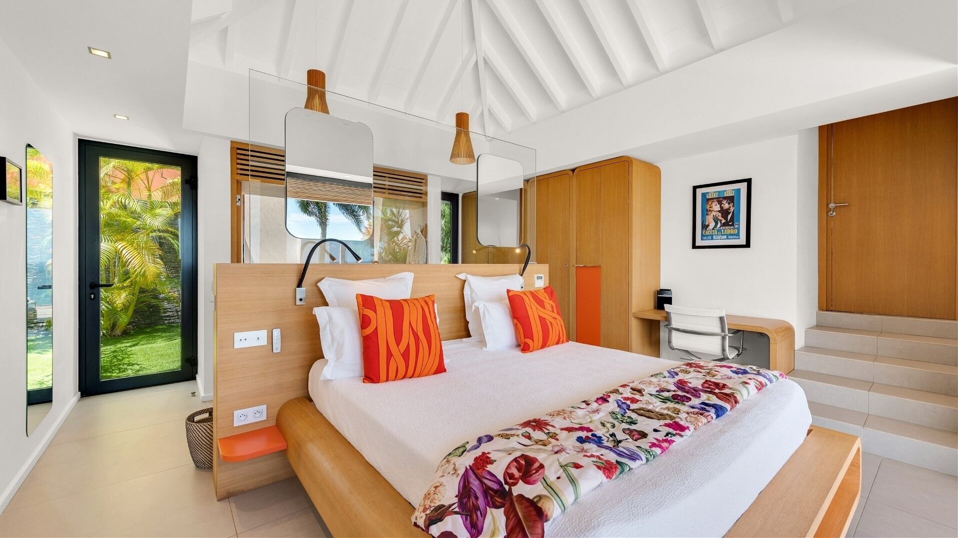 Une chambre moderne avec un grand lit, une tête de lit en bois, un bureau et une vue sur des palmiers, visible à travers une porte vitrée.