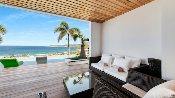Une pièce moderne avec un plafond en bois, une table en verre, un canapé en osier avec des coussins blancs, et une vue sur la plage et l’océan.