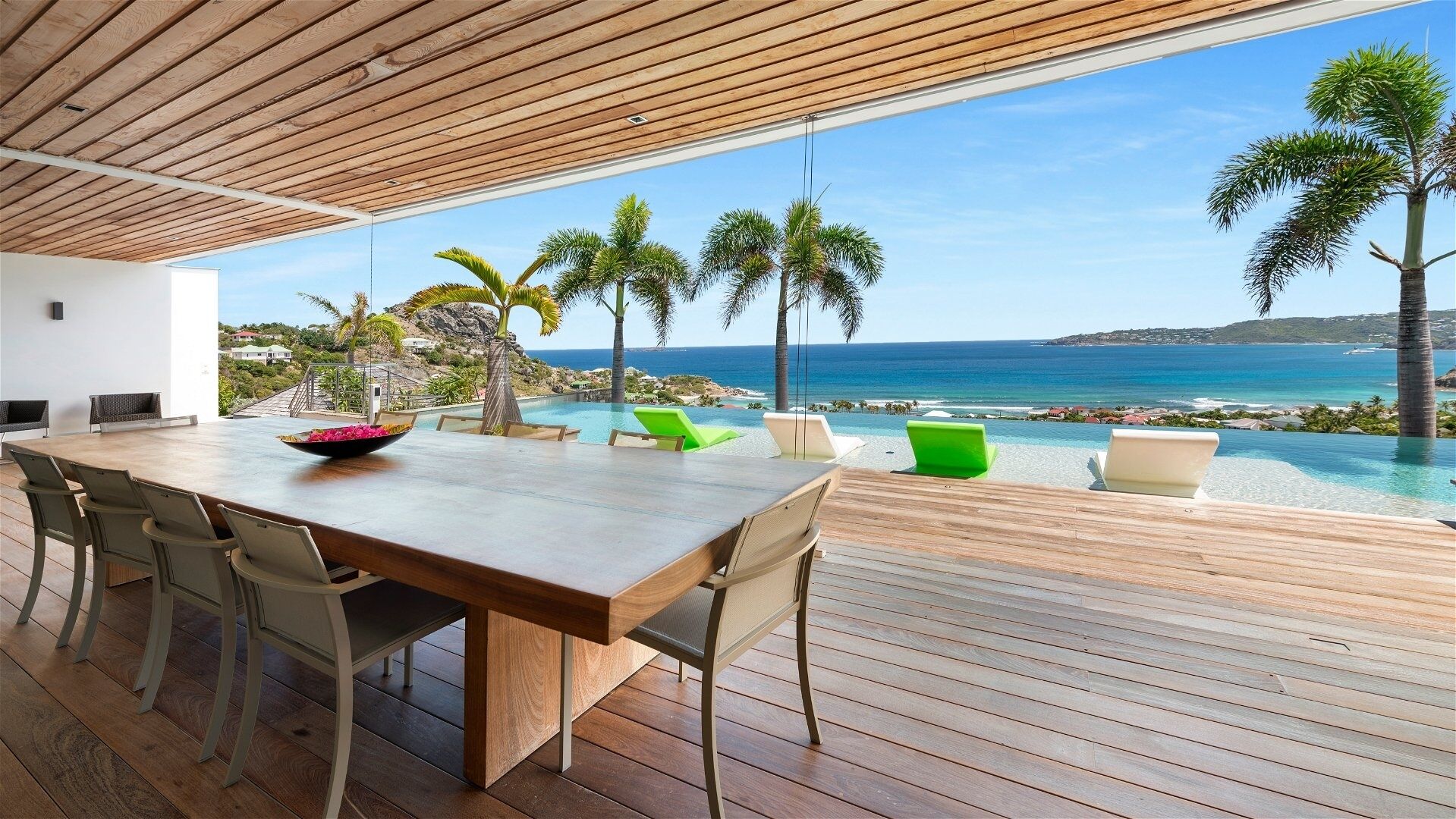 A wooden deck with a dining table and chairs, overlooking a pool and the ocean.