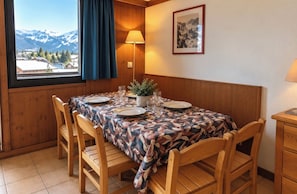 A dining area with a table set for four, wooden chairs, a view of mountains through a window, and a lamp on the wall.