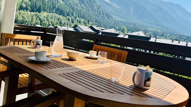 A wooden table set with a mug, glass, and bowl on a balcony overlooking a mountain range. - Nice flat with balcony view Mont Blanc | Parking (Chamonix-Mont-Blanc)