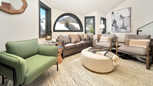 A modern living room with a green sofa, a brown couch, and a round ottoman. There are two windows with arched tops, a lamp, and a painting on the wall. - Lulu 6J Walk to Lift 7 + Pool & HotTub - 2 Bedroom+Loft, 3 Bathroom, Sleeps 6 (Telluride)
