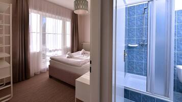 A hotel room with a bed, a bedside table, a wardrobe, a shower area with blue tiles, and a large window with sheer curtains.