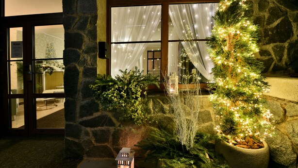 Een stenen muur, een kerstboom in pot met lichtjes, en een verlichte lantaarn.