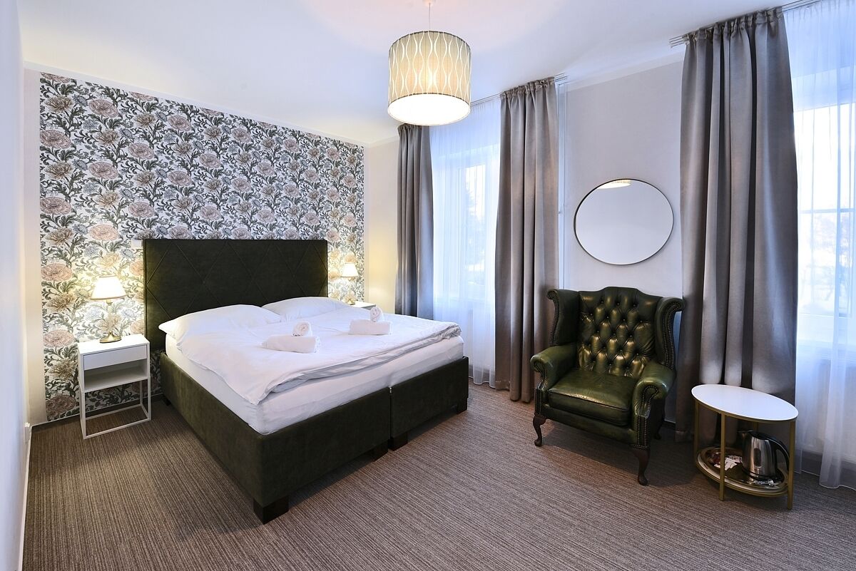 A hotel room with a large bed, a green armchair, a round mirror, and a small table with a kettle.