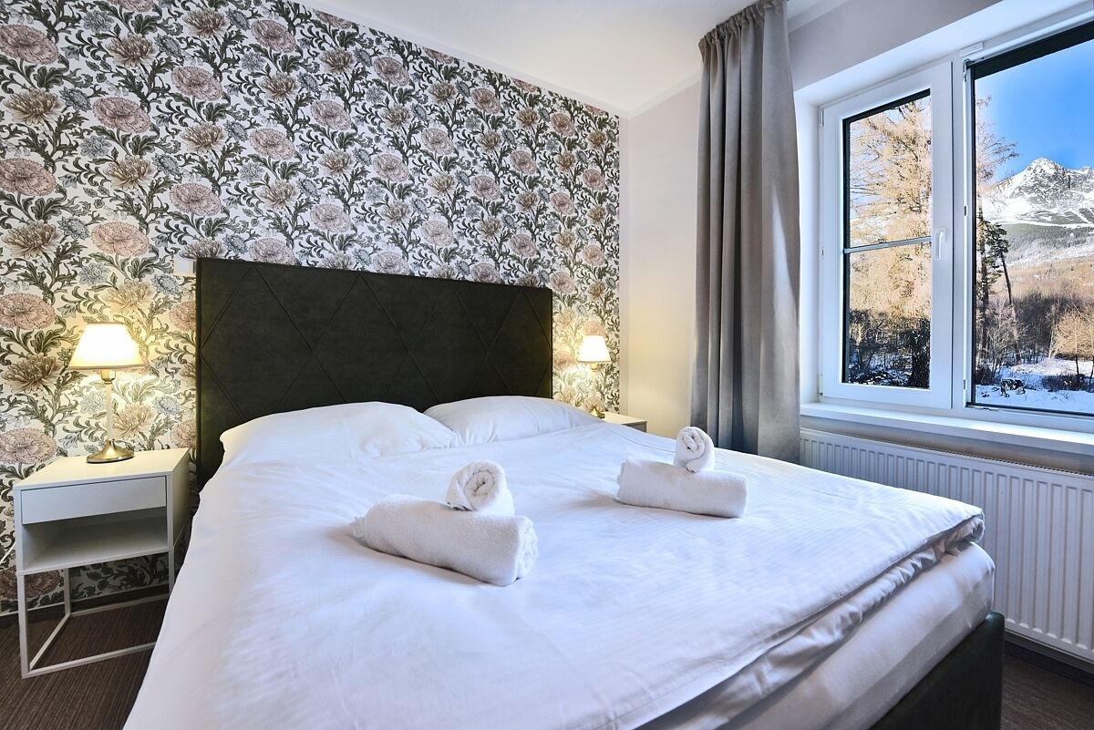 A hotel room with a large bed, floral wallpaper, two bedside lamps, and a window with a snowy mountain view.