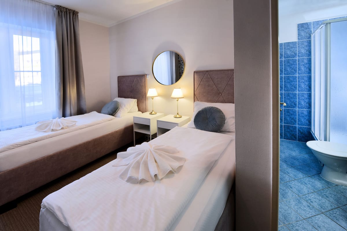 A hotel room with two beds, a round mirror, bedside lamps, and a bathroom with a blue-tiled shower.