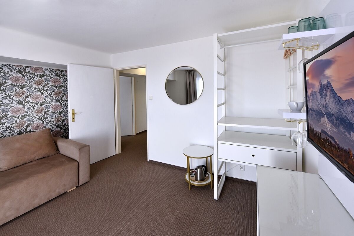 A hotel room with a sofa, a round mirror, a small table with a kettle, a TV, and a bookshelf.