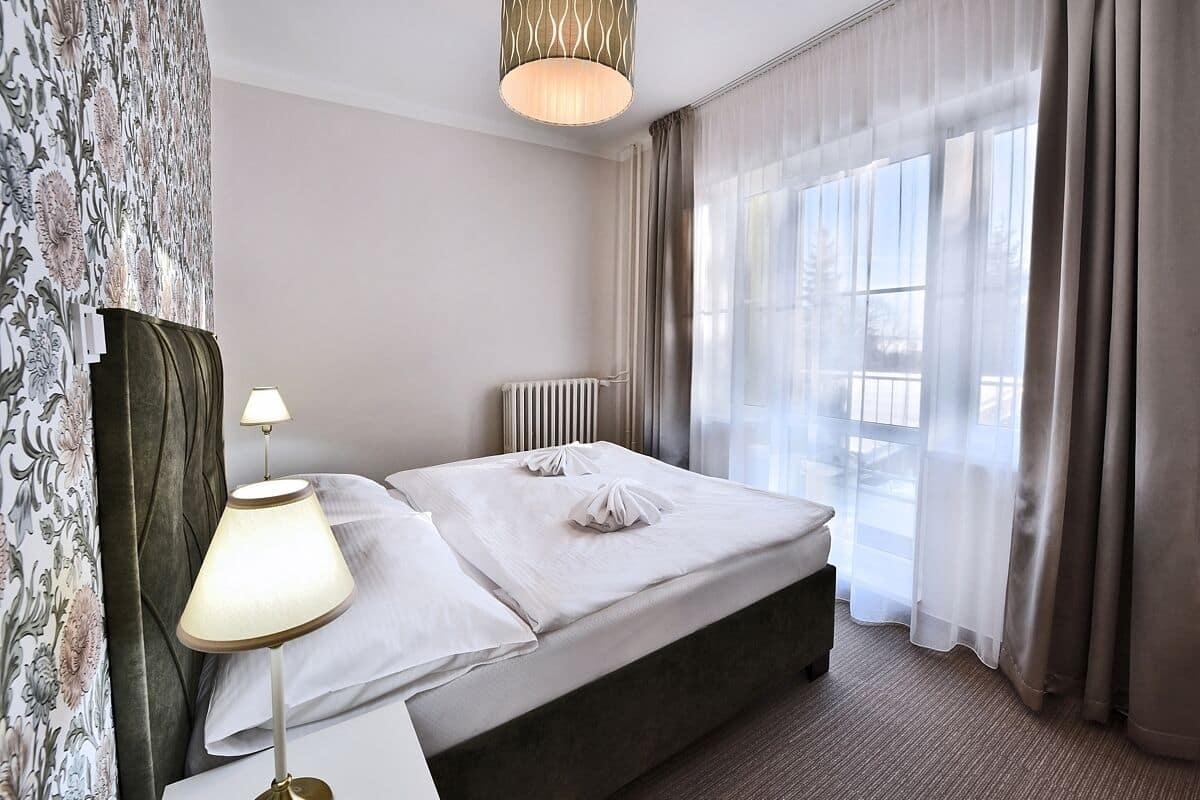 A hotel room with a neatly made bed, a bedside lamp, a radiator, and a large window with curtains.