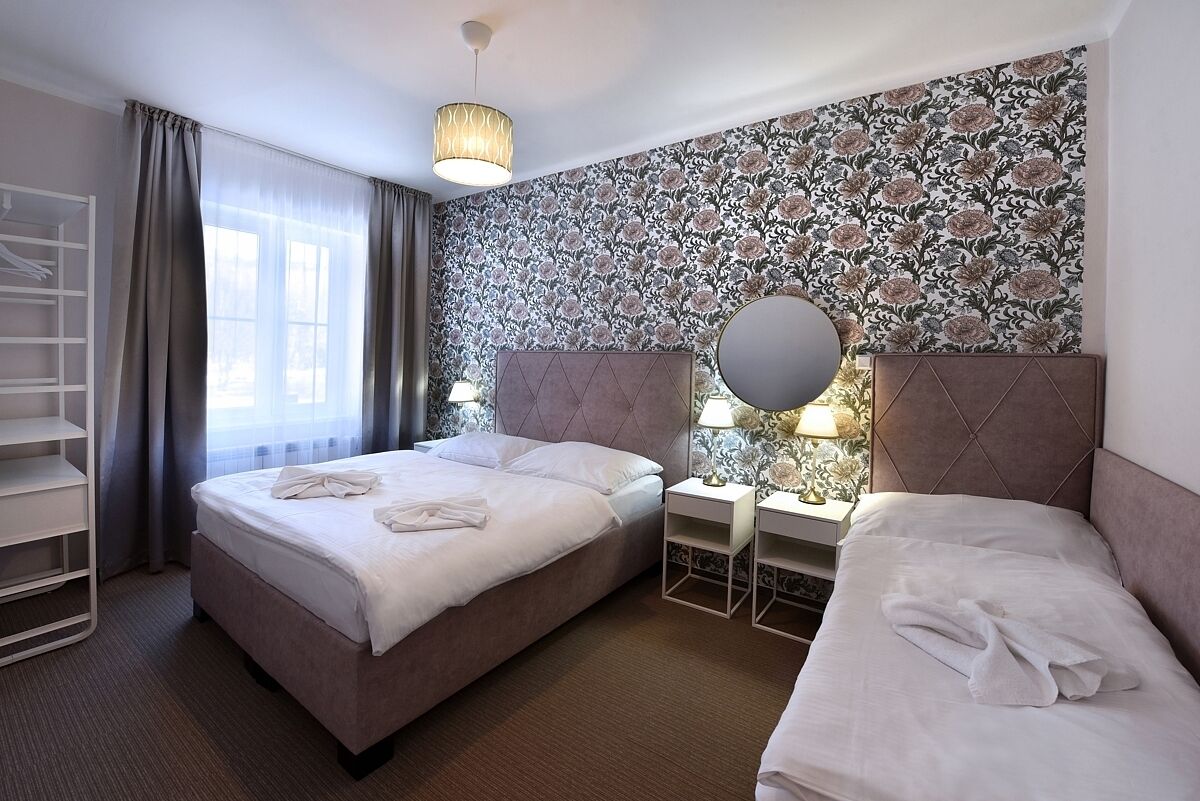 A hotel room with two beds, a wardrobe, a large window with curtains, a floral wallpaper, and a round mirror on the wall.