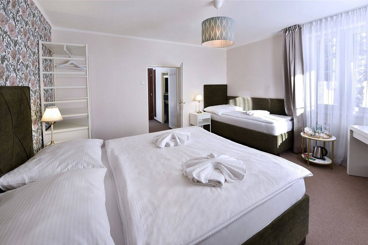 A hotel room with two beds, a wardrobe, a bedside table with a lamp, a small table with cups and a kettle, and a view of the city through sheer curtains.