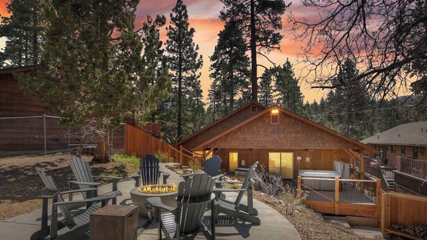 Une cabane dotée d’un foyer et de sièges extérieurs, entourée de grands arbres et baignée par les couleurs du coucher de soleil.