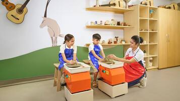 Un aula con niños creando cerámica, rodeada de estanterías con artículos de alfarería y una guitarra en la pared.