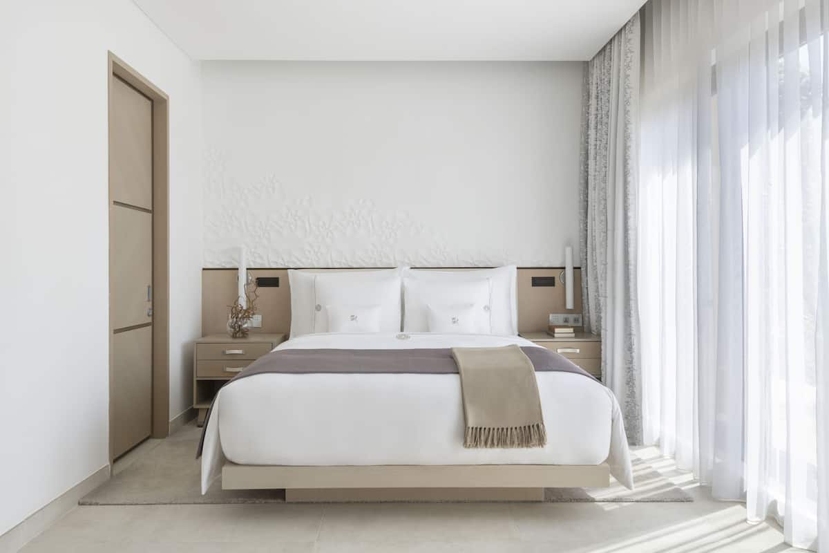 Une chambre d’hôtel moderne, dotée d’un grand lit, de tables de chevet et offrant une vue sur l’extérieur à travers de fines rideaux.