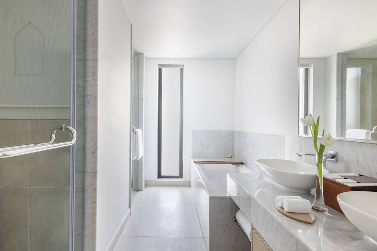 Une salle de bain moderne avec une douche à l’italienne en verre, un grand miroir et un lavabo blanc agrémenté d’un vase de fleurs.