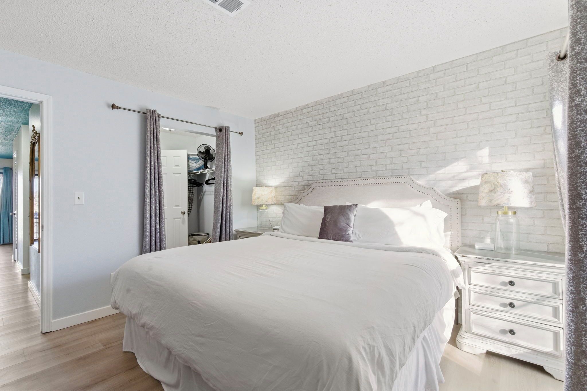 A bedroom with a large bed, a white brick wall, a nightstand with a lamp, and a door leading to a bathroom.