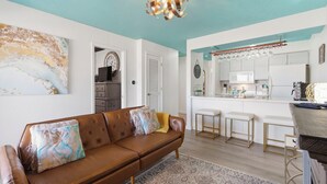 Una sala de estar con un sofá de cuero marrón, un tapete estampado y una zona de cocina con armarios blancos y una barra.
