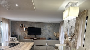 A modern living room with a wooden dining table, a flat-screen TV mounted on the wall, and a sofa with patterned cushions. - Villa design swimming pool Avignon festival (Avignon)