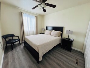 A bedroom with a bed, a chair, a nightstand, a ceiling fan, and a window with curtains.