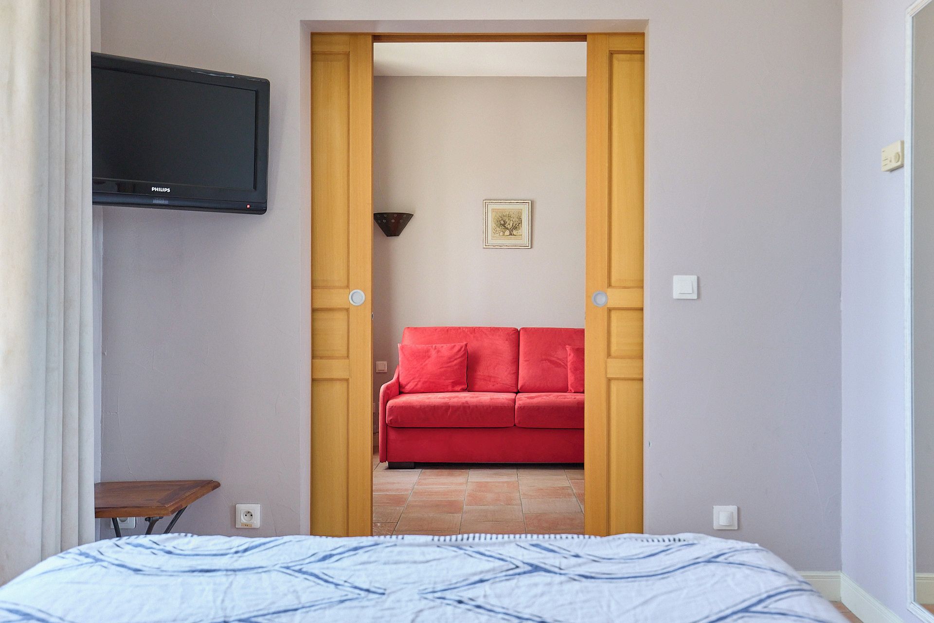 A bedroom with a bed, a television mounted on the wall, a red sofa in the adjacent room, and a door leading to another space.