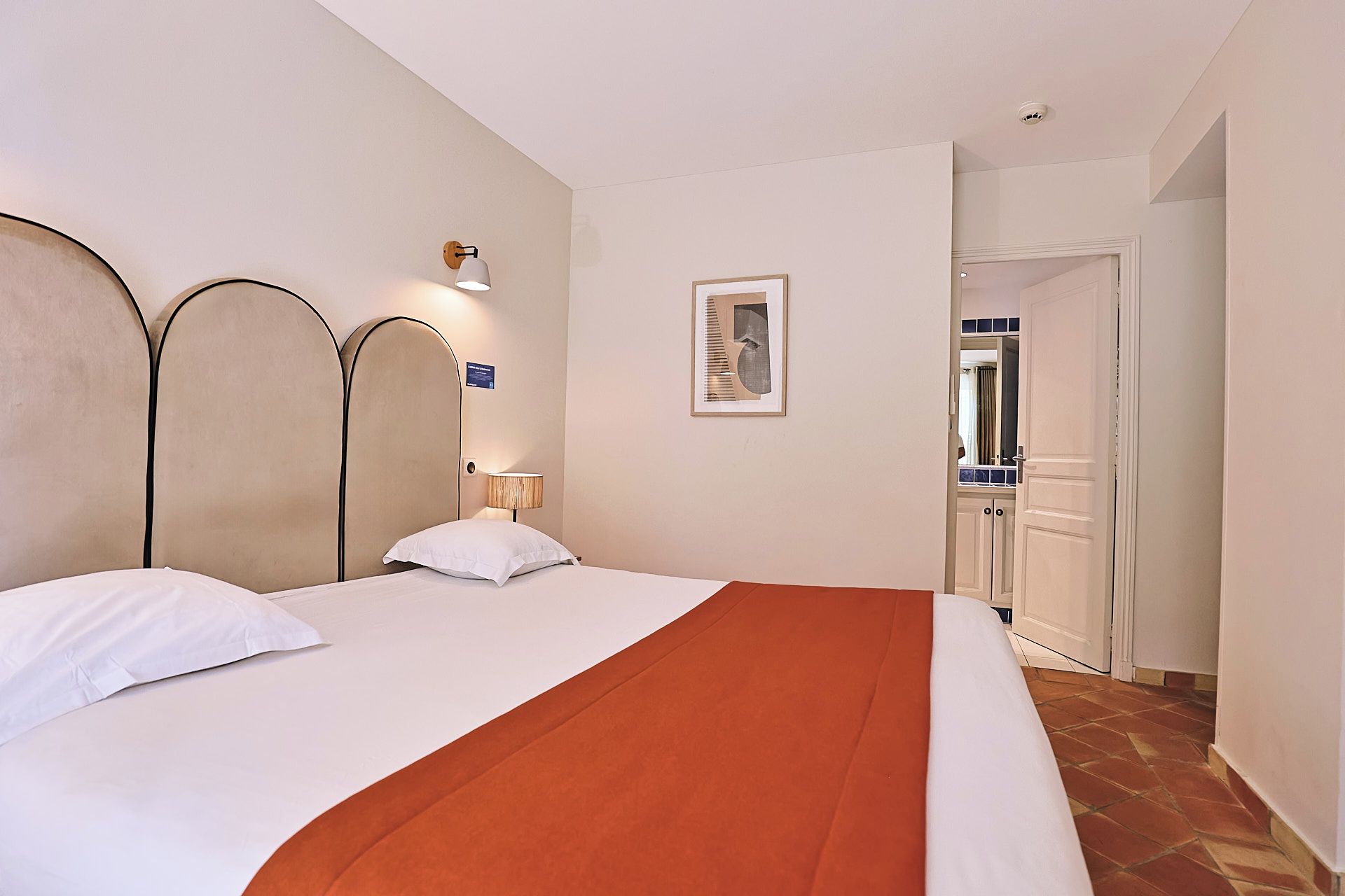 A hotel room with a bed, a wall-mounted lamp, a framed picture, and a door leading to another room.