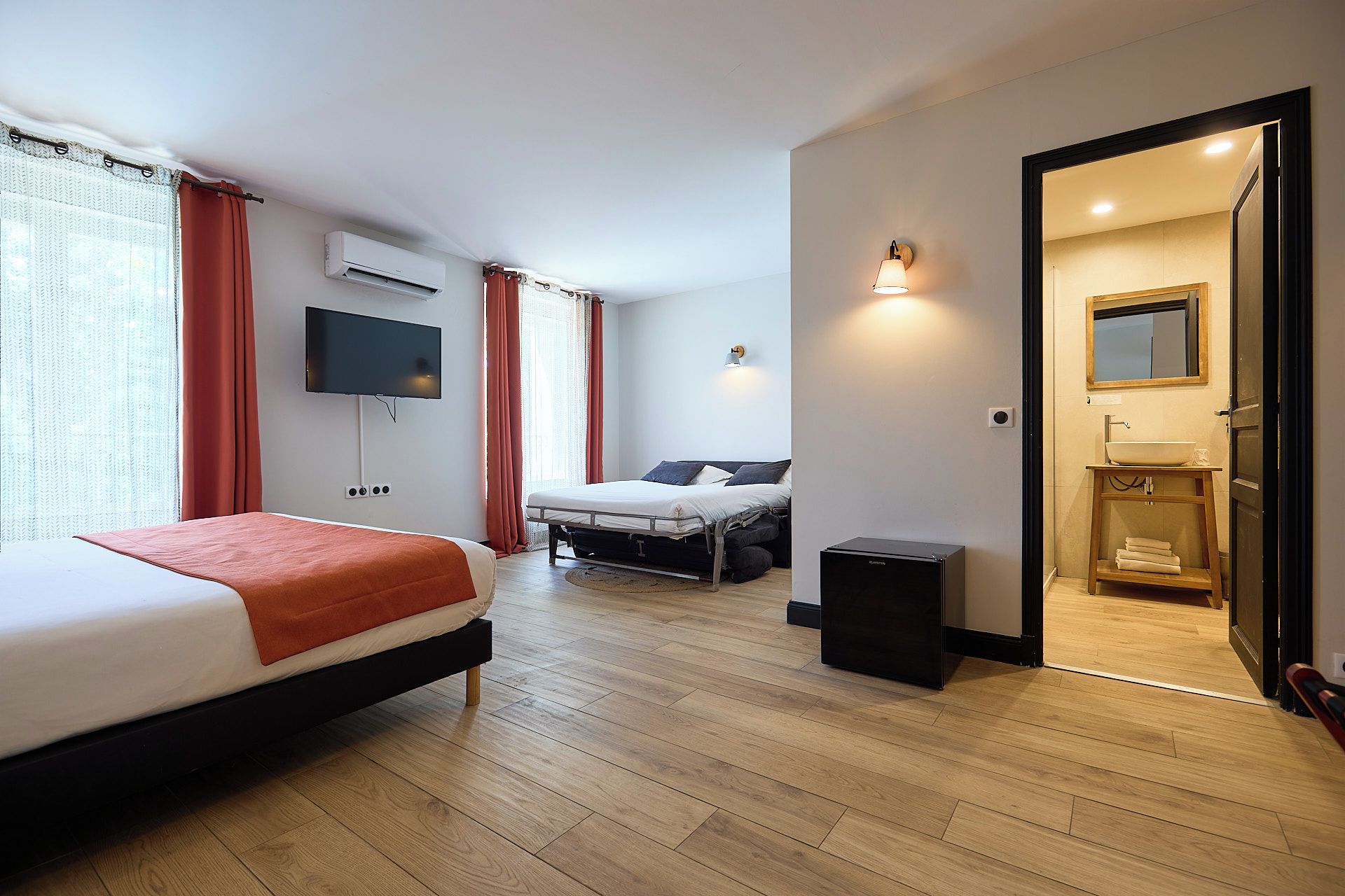 Habitación de hotel con cama, televisión, mesita de noche, un taburete pequeño y un baño visible a través de una puerta abierta.