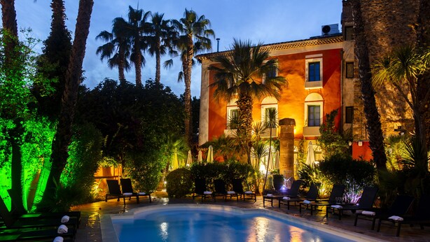 A swimming pool surrounded by lounge chairs and palm trees, with a building in the background. - Hôtel l'Arena (Fréjus)