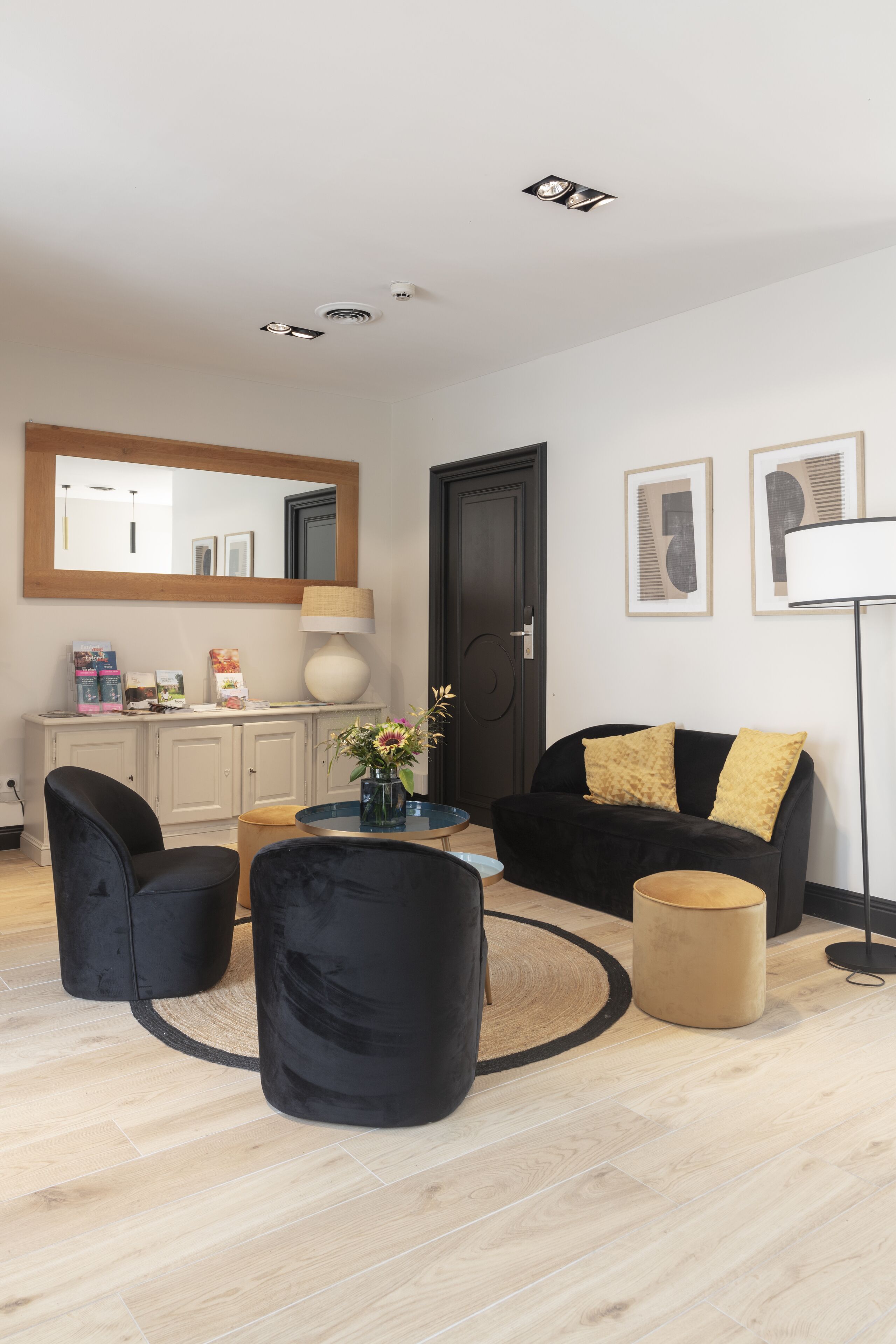 A modern living room with a black sofa, two black armchairs, a round coffee table, and a beige ottoman. There is a large mirror above a white cabinet, a floor lamp, and framed artwork on the wall.