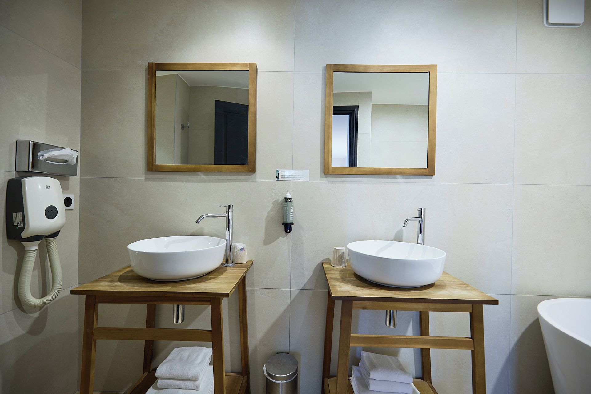 A bathroom with two sinks, wooden vanity tables, a hand dryer, and a trash can.