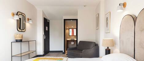 Quarto de hotel com cama, cadeira, escrivaninha com luminária e espelho fixado na parede.