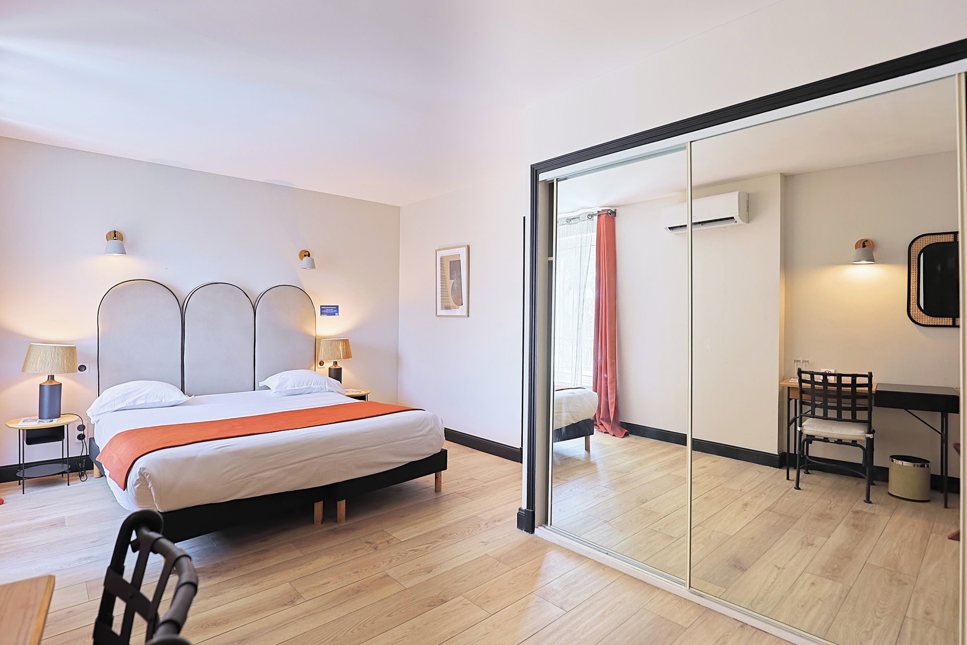 Une chambre d’hôtel comprenant un lit, des lampes de chevet, un bureau avec une chaise, un miroir et une porte-fenêtre coulissante donnant sur une salle de bain.
