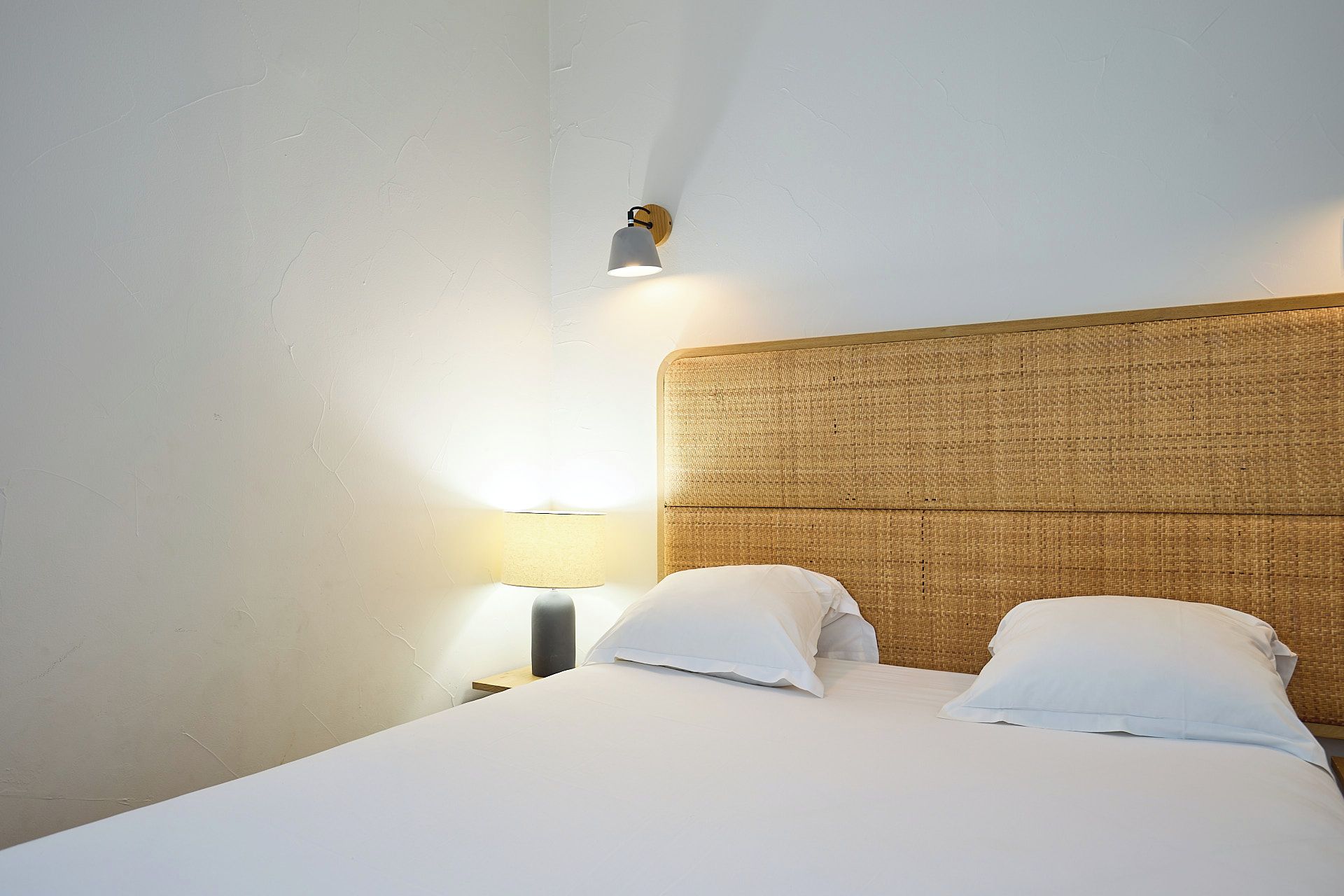 A hotel room with a bed, two pillows, a nightstand with a lamp, and a wall-mounted light fixture.