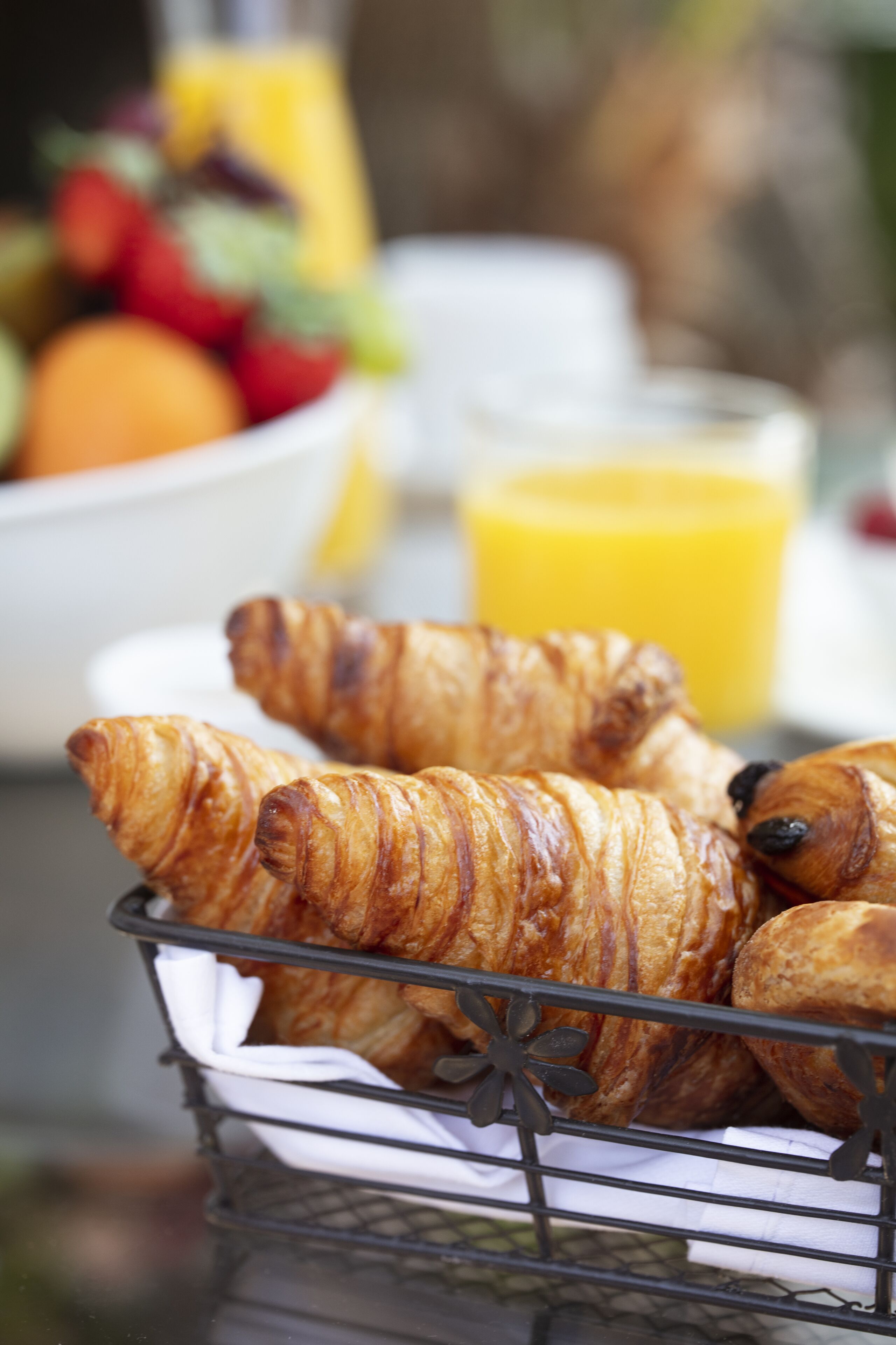 A basket of croissants on a table with a bowl of fruit and a glass of orange juice in the background.