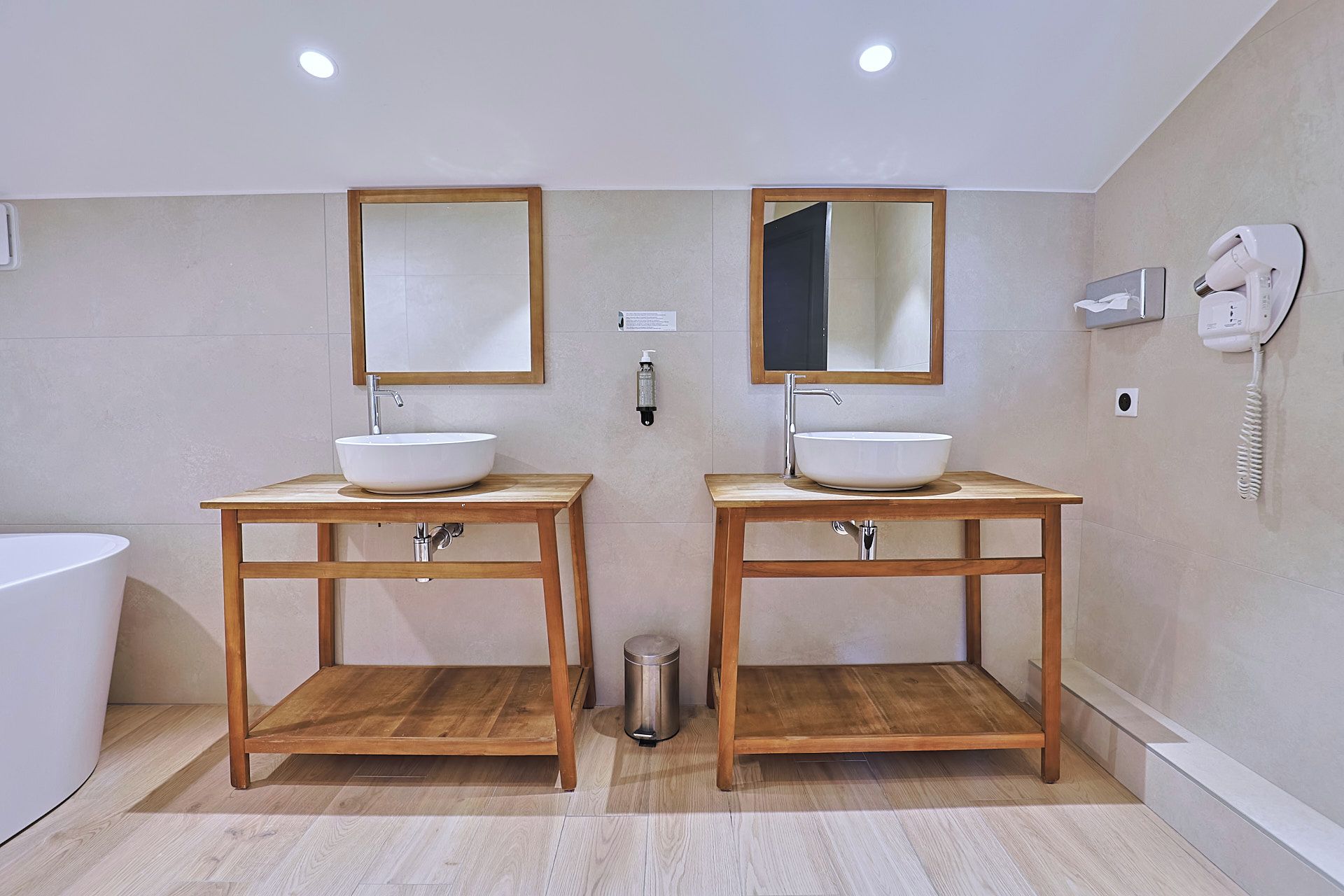 A bathroom with two wooden vanity tables, each with a white sink and a mirror above. There is a hairdryer on the wall to the right.